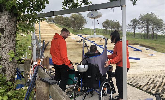 Two volunteers in orange jackets help a man in a grey sling be hoist transferred from wheelchair to sit ski.